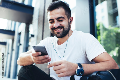 Cheerful hipster guy in electronic headphones using smartphone technology with media application for listening online radio, happy man in earphones enjoying positive music while chatting in networks