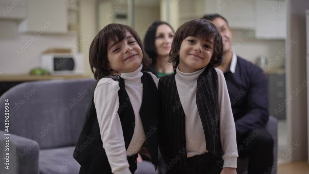 Twin brothers hugging smiling looking at camera with blurred parents resting at background watching TV. Positive Middle Eastern children posing indoors at home Twin brothers hugging smiling looking at camera with blurred parents resting at background watching TV. Positive Middle Eastern children posing indoors at home