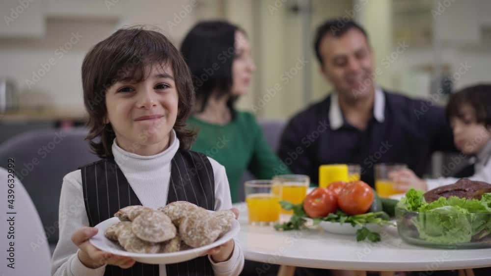 Smiling cute Middle Eastern boy posing with delicious bakery at home with blurred family sitting at table at background. Happy son having dinner with twin brother mother and father Smiling cute Middle Eastern boy posing with delicious bakery at home with blurred family sitting at table at background. Happy son having dinner with twin brother mother and father