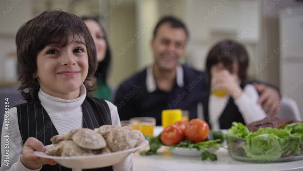 Portrait of cute boy holding plate with sweet delicious bakery looking at camera as blurred family enjoying dinner at background. Happy smiling Middle Eastern child posing indoors Portrait of cute boy holding plate with sweet delicious bakery looking at camera as blurred family enjoying dinner at background. Happy smiling Middle Eastern child posing indoors