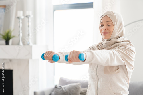 Healthy muslim islamic arabian woman exercising at home with dumbbells in hijab, fitness and workout on lockdown. Keeping fit body being in shape