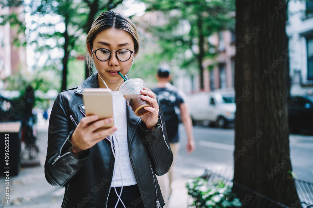 Millennial travel blogger in optical eyewear and headphones listening received audio message during touristic vacations, Chinese hipster girl drinking takeaway beverage and choosing music records Millennial travel blogger in optical eyewear and headphones listening received audio message during touristic vacations, Chinese hipster girl drinking takeaway beverage and choosing music records