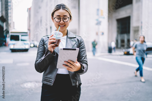 Happy Asian blogger with takeaway coffee watching content video browsed on modern touch pad, cheerful hipster girl using digital tablet for networking and reading publication in social media