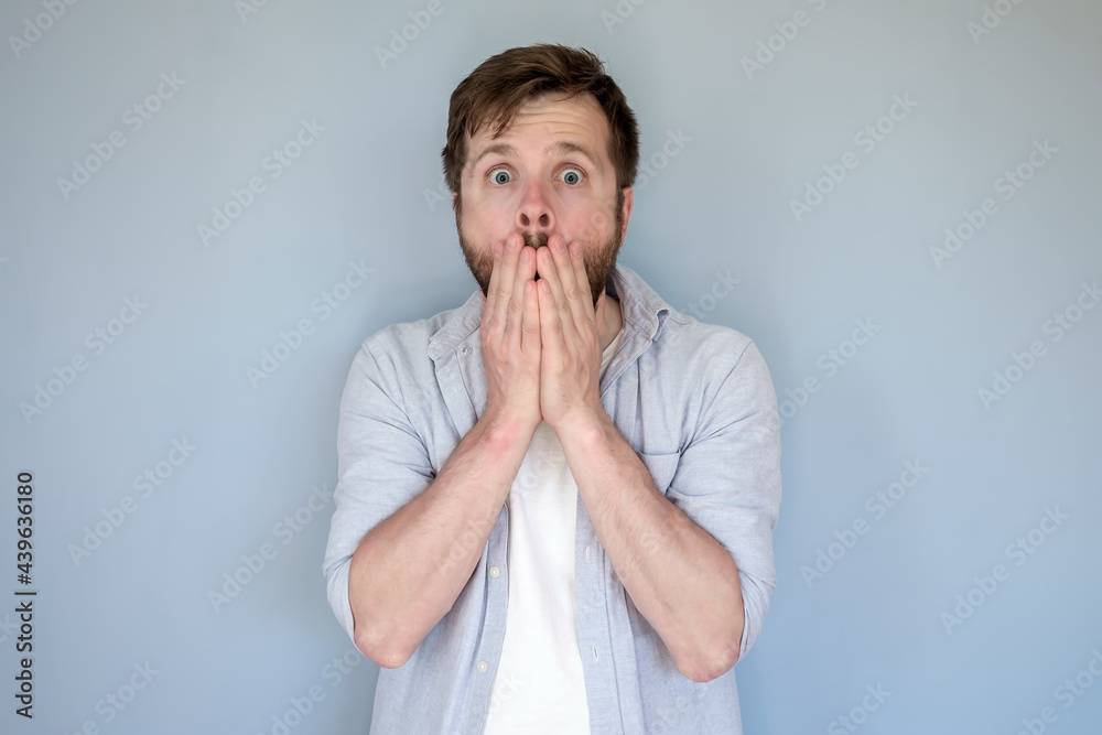 Shocked man in casual clothes puts hands over his mouth in fear and looks at the camera with big eyes. Gray background. Shocked man in casual clothes puts hands over his mouth in fear and looks at the camera with big eyes. Gray background.