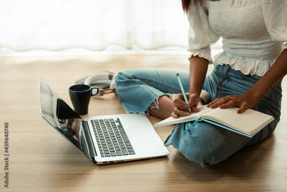 Naklejka premium Close-up of a woman sitting on the floor holding a pencil and taking notes using a laptop at home.