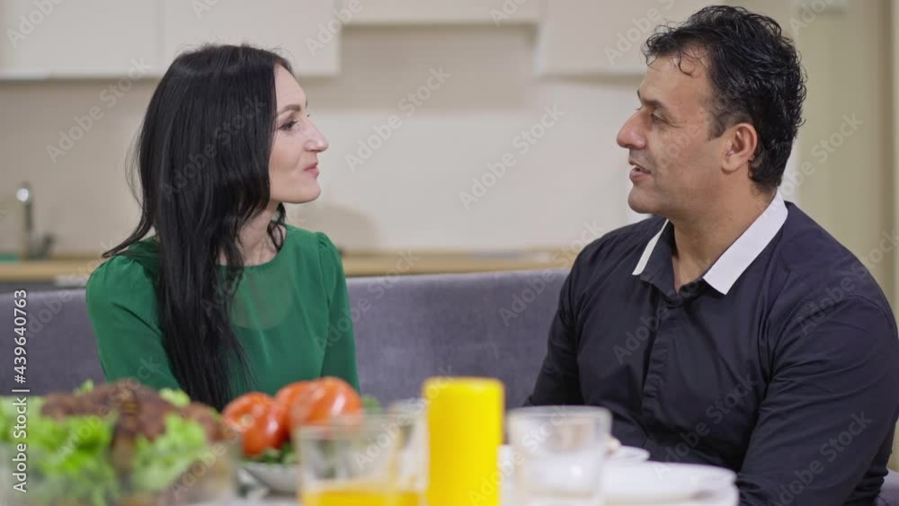 Side view portrait of happy loving interracial couple talking and smiling discussing day in the evening at home indoors. Adult Caucasian woman and Middle Eastern man chatting resting at dinner Side view portrait of happy loving interracial couple talking and smiling discussing day in the evening at home indoors. Adult Caucasian woman and Middle Eastern man chatting resting at dinner