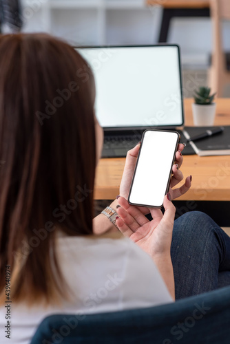Back view of woman holding a smartphone blank white screen sitting at the office. Mock up.