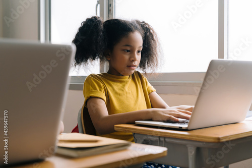 Cute little African American girl with curly hair and ponytail in school sitting in classroom studying while typing on laptop with focus and concentration