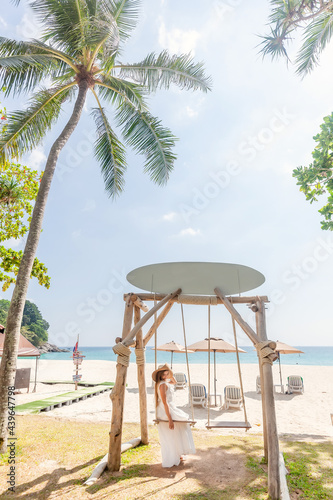 Travel Woman in White Dress and Straw Hat Sitting on Wooden Swing, Tropical Palms and Sandy Beach with Blue Sea on Background. Female Tourist Leisure in Resort on Phuket Island, Thailand © TravelMedia