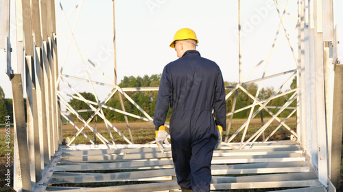 An electrician or builder and engineer, in a blue robe, in a yellow and white helmet, check the technical overview for strength, prepare electric poles, an electric tower, energy, and workers. 