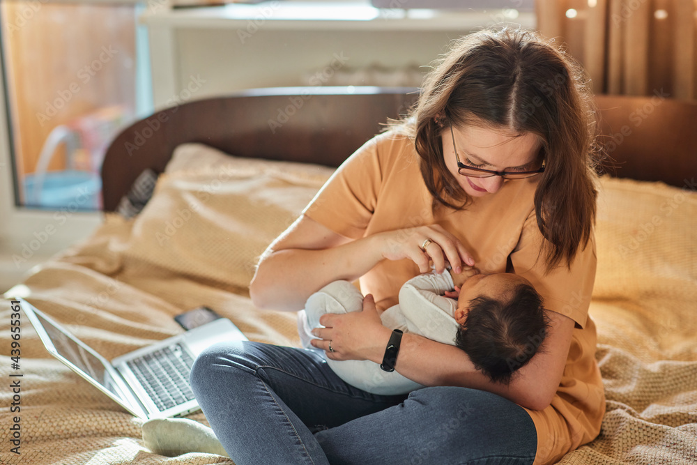 Mother multi-tasking, holding baby infant and using computer laptop at home. 
