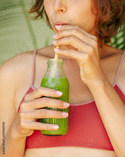 Portrait of a young girl in a swimsuit with a bottle of green smoothie near palm leaves 