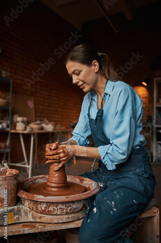 Beautiful positive craft woman wearing apron making clay pot on pottery wheel in workshop photo