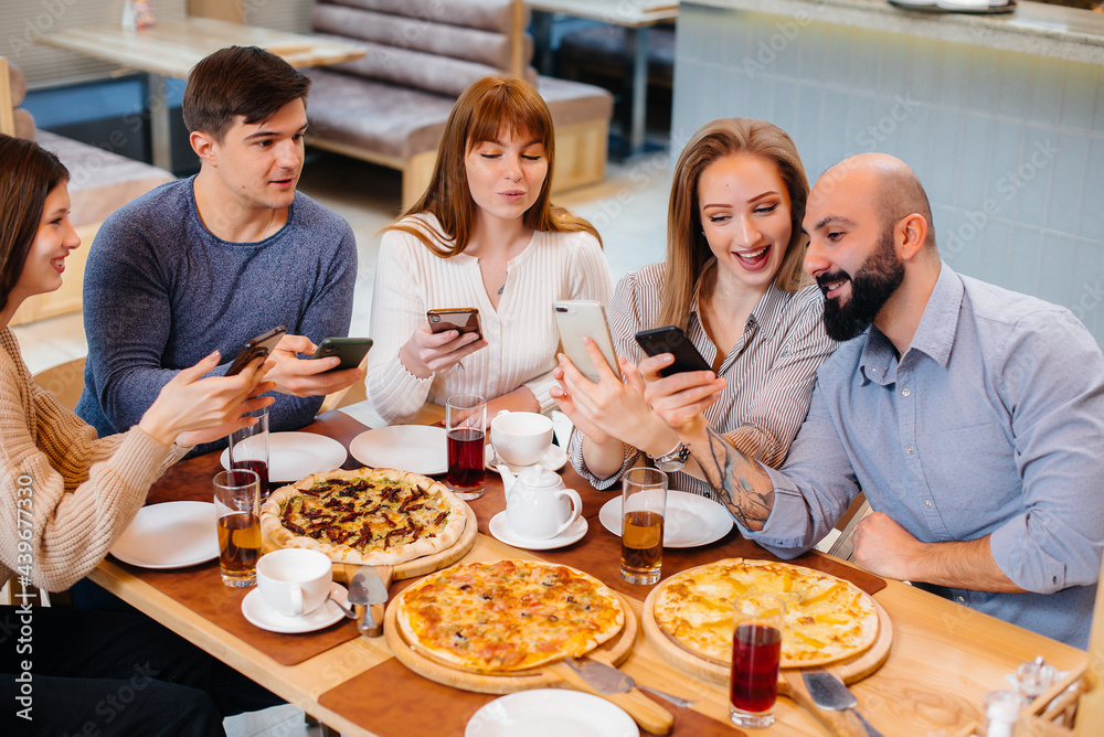 A group of young cheerful friends is sitting in a cafe talking and taking selfies on the phone. Lunch at the pizzeria. A group of young cheerful friends is sitting in a cafe talking and taking selfies on the phone. Lunch at the pizzeria.