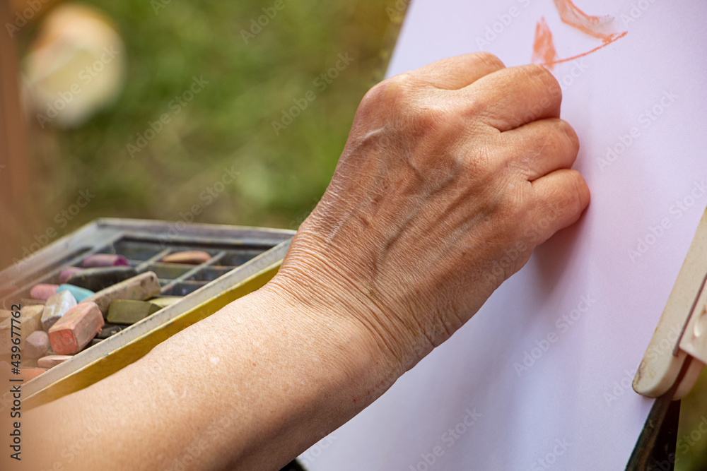 Street artist holding a box with multicolored crayons and pencils for drawing.Close up photography of hands and drawing supplies.He drawing behind the easel with white paper.
