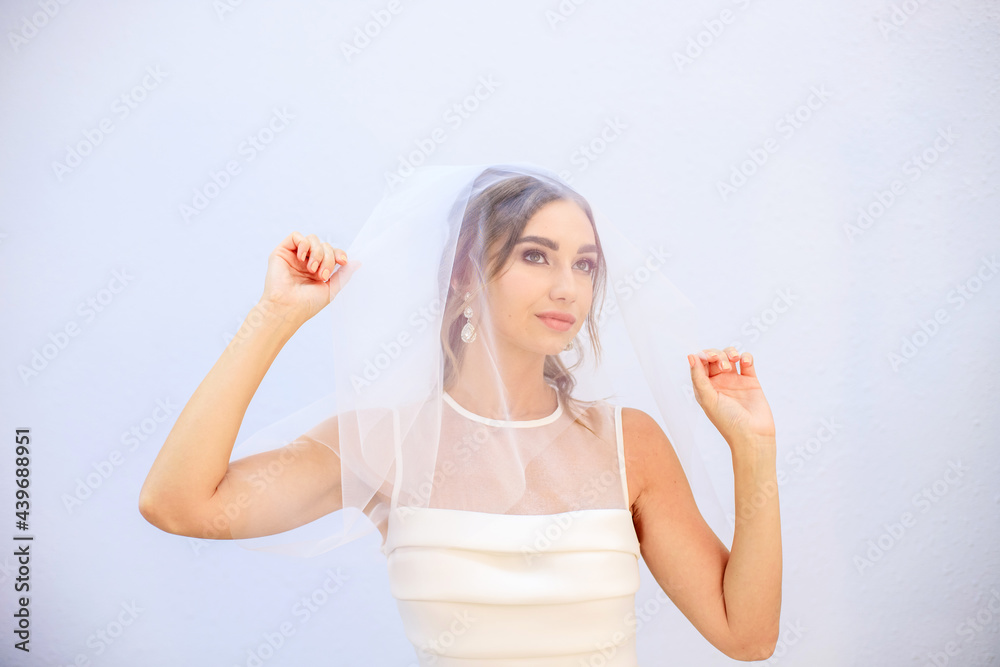 A brunette girl in a wedding dress covers her face with a veil. Bride in a white dress. Makeup. A brunette girl in a wedding dress covers her face with a veil. Bride in a white dress. Makeup.