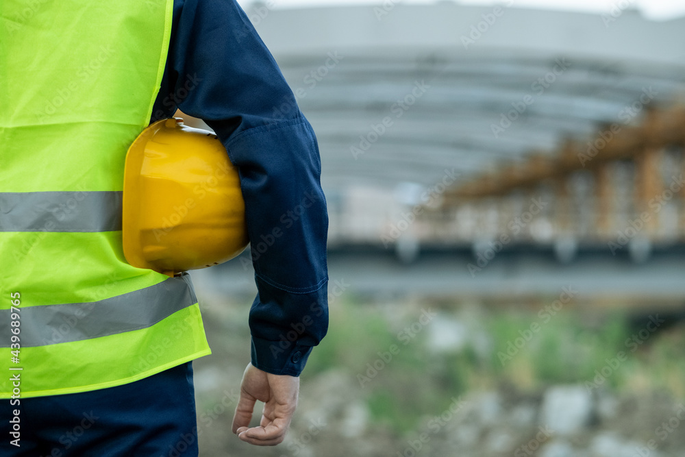 Rear view of contemporary builder holding helmet under left arm Rear view of contemporary builder holding helmet under left arm