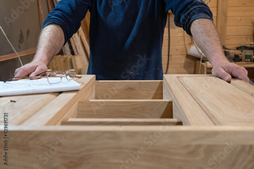 hands of a master carpenter who makes a handmade cabinet made of natural wood in light colors. Next to the glasses and the drawing. Production of custom-made furniture