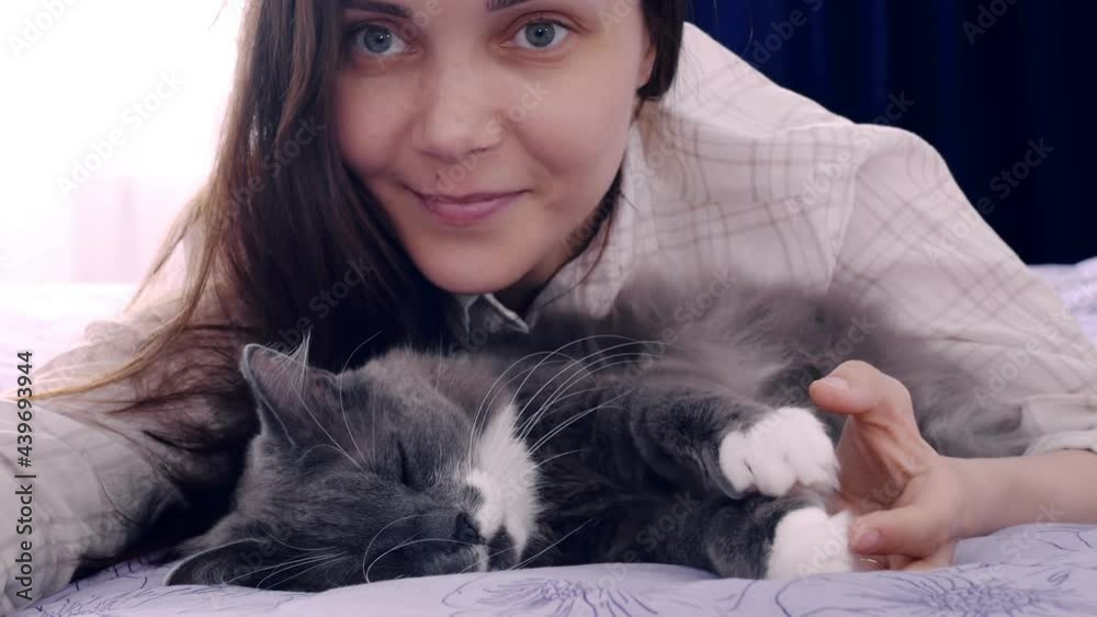 A dark-haired woman takes pictures of herself and her fluffy cat on a smartphone. The woman smiles sweetly, the pet sleeps wiggling its paws in a dream. Selfie with animals on a smartphone.
