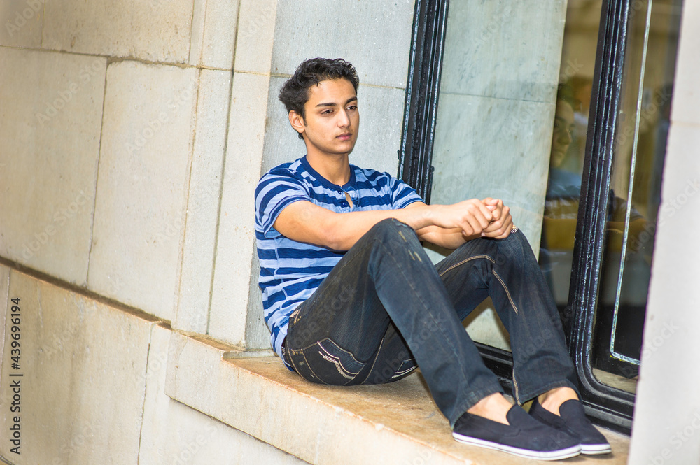 Dressing in a blue lines T shirt and  black pants, a young asian teenager is sitting on a window and into deeply thinking.