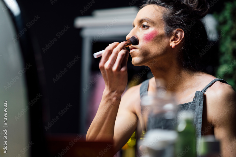 Side view of male gay sitting at table and doing makeup in drag queen style while applying powder with brush Side view of male gay sitting at table and doing makeup in drag queen style while applying powder with brush