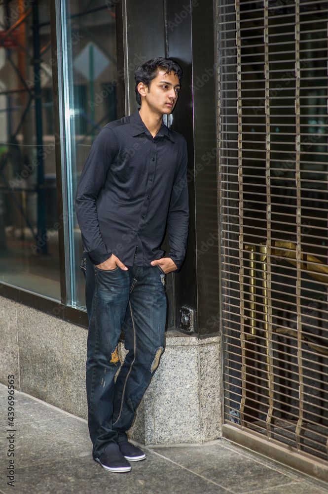 Dressing in a black shirt and a fashionable pants, a young asian teenager is leaning on a closed door, looking inside and thinking.