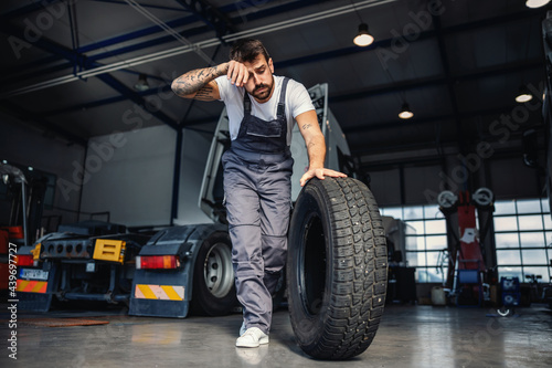 Tired hardworking mechanic rolling tire in order to change it on truck. He is in garage of import and export firm. © dusanpetkovic1