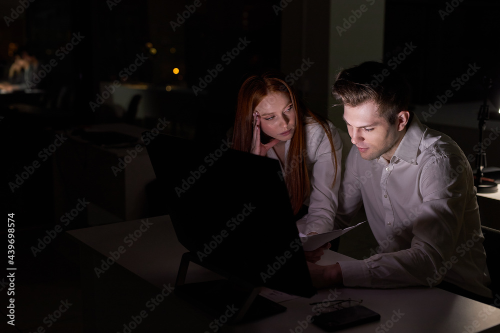 Two young caucasian business people working together on computer, discussing project in office at late night. side view on redhead lady and handsome guy colleagues brainsotrming. in dark boardroom