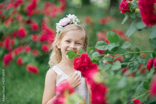 Summer portrait of pretty little girl wearing white dress and hat, posing in rose garden