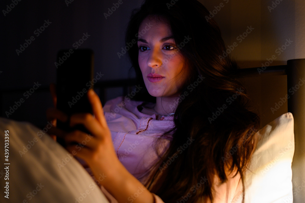 Young beautiful woman lying in her bed at night and using smartphone