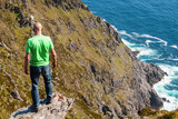 Bald male tourist standing on the edge of a cliff, Beautiful scenery in the background. Warm sunny day, Achill island, county Mayo, Ireland. Adventure and risk and reckless behavior concept