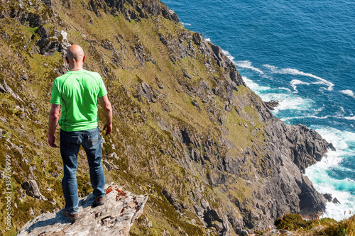 Bald male tourist standing on the edge of a cliff, Beautiful scenery in the background. Warm sunny day, Achill island, county Mayo, Ireland. Adventure and risk and reckless behavior concept photo