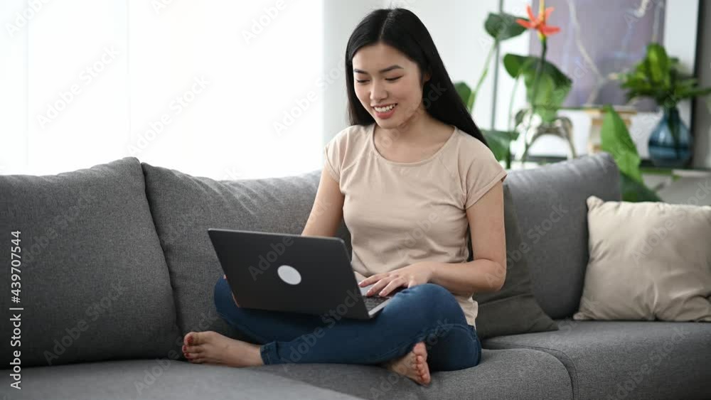 Video communication, online conference. Young pretty asian woman, freelancer or student, in casual wear, sits on a sofa at home, uses laptop to communicate with friends on a video call, talks online Video communication, online conference. Young pretty asian woman, freelancer or student, in casual wear, sits on a sofa at home, uses laptop to communicate with friends on a video call, talks online