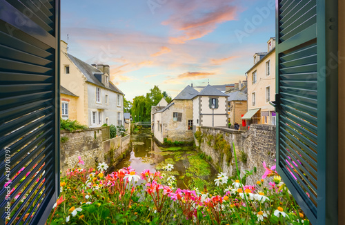 View through an open window with shutters above a small stream with medieval homes nearby, at sunset in the French Village of Bayeux, France, in the Normandy region.