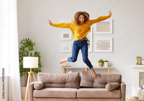 Fototapeta Naklejka Na Ścianę i Meble -  Happy african american teen girl jumping on sofa while having fun on weekend at home