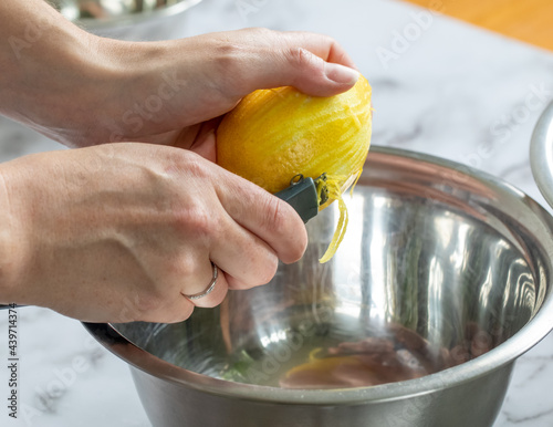Deux mains féminines en train de peler la pelure d'un citron jaune pour en extraire le zeste au dessus d'un bol à mélanger en métal sur lumière naturelle photo