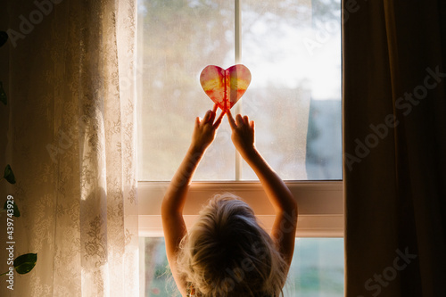 A little girl putting a paper heart against the window at the sunrise photo