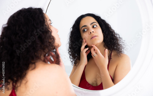 Young focused woman touching face and looking at reflection in mirror in bedroom