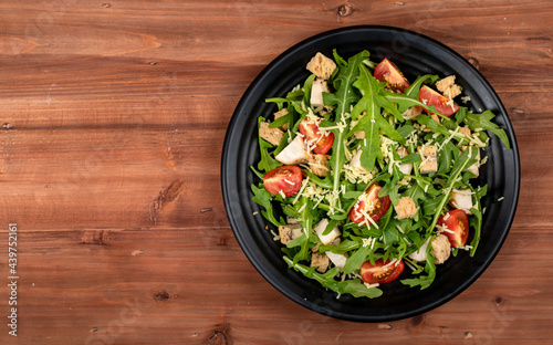 Fresh rocket salad with tomato, crispy crouton, small pieces of herb roasted chicken and parmesan cheese in black ceramic dish on the right side of dark wooded table