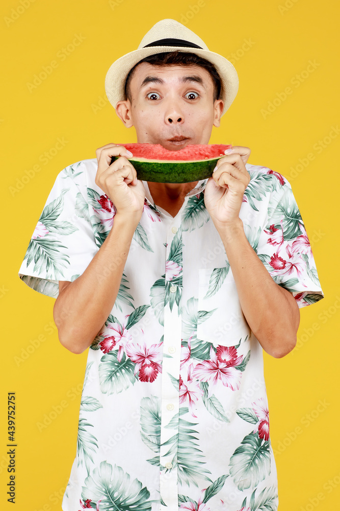 Young attractive Asian man in white Hawaiian shirt wearing white hat eating watermelon slice with excitement against yellow background. Concept for beach vacation holiday
