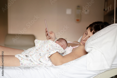 Mother lying in a bed with her baby on top of her using a mobile