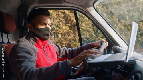 Indian man looking at the laptop placed on the dashboard of the car. Dressed in red upper wearing a black mask sitting on the driving seat of the car using a laptop placed on the dashboard of the car.