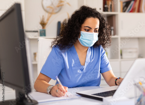 Portrait of female doctor wearing protective mask working on laptop computer consulting patient online, telemedicine concept