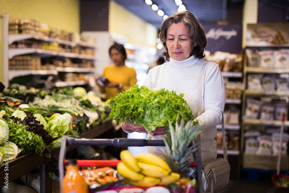 Female elderly shopper choosing ripe salad at grocery supermarket