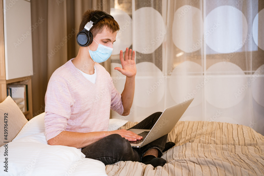 a young man of european appearance in casual clothes sits on a bed in a room at home wearing headphones and a protective mask at home with a laptop, the concept of remote work and education