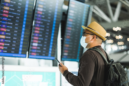 Young traveler man wearing mask check in Arrival or departure passenger standing at information Flight schedule on dashboard. Tourist in airport terminal. Backpacker travel in downtown urban city.