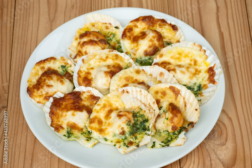 Baked parmesan scallops in a white ceramic plate on a wooden table . Oven baked scallop with cheese  spinach  butter and garlic.