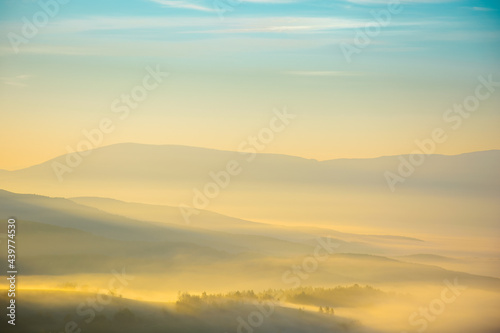 mountainous countryside landscape at foggy sunrise. wonderful autumnal nature scenery with distant rural valley in glowing mist. trees and fields on hills in morning light