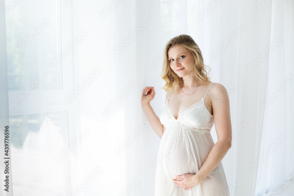 Happy blonde pregnant woman at the window in the morning in a beautiful negligee Happy blonde pregnant woman at the window in the morning in a beautiful negligee