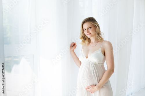 Happy blonde pregnant woman at the window in the morning in a beautiful negligee